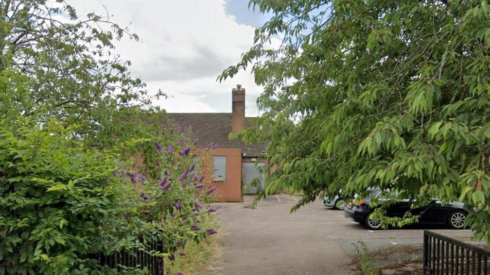 An old school building, including cars parked in the car park, trees, and bushes. The school building is red brick.