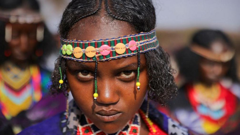 A young girl with a beaded headband looks directly into the camera during a swearing ceremony in Arero, Ethiopia - Sunday 9 March 2025.