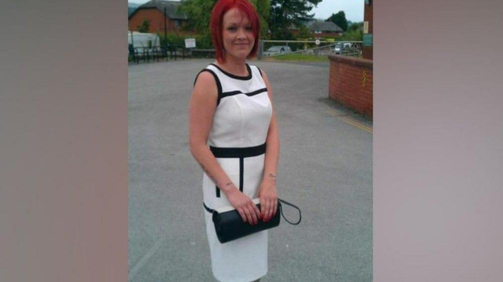 Tiffany Render has dyed-red hair and is smiling. She is standing near a building, dressed in a smart long white and black dress holding a black handbag. 