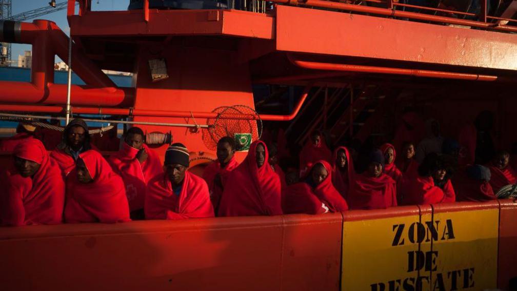 Migrants and refugees wearing blankets to stay warm are standing on a boat, operated by Spain's Maritime Rescue Service