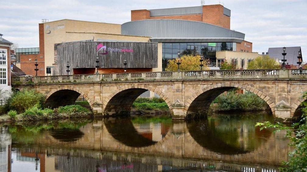 A bridge across the River Severn, with a modern theatre building to the side of the river reflected in the water