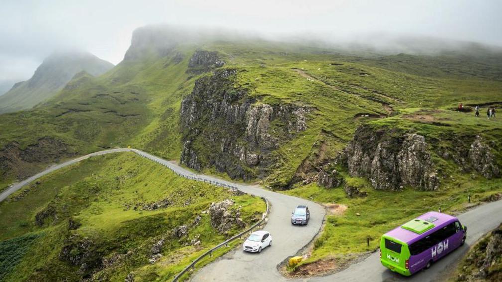 Two cars and a green and purple tour bus drive along a winding road at the Quiraing on Skye. The rocky landscape of the Quiraing is shrouded in low cloud and mist.