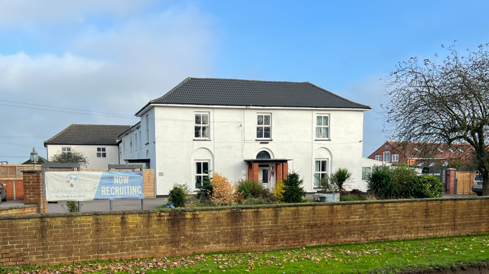 Large white, Georgian-looking care home in background with a wing to the side and a car park to the front. A waist-high brick wall separates the building from the verge and a road. 