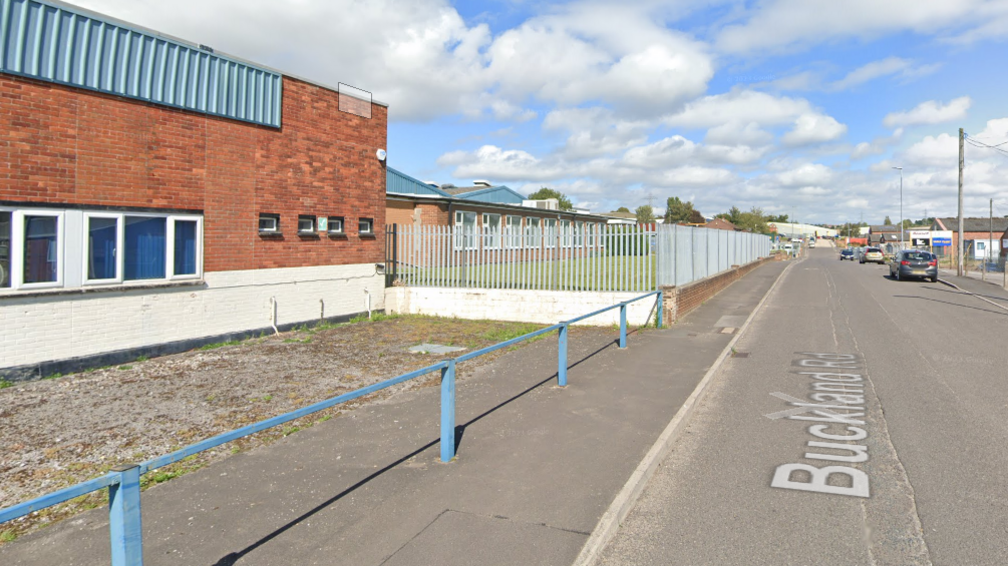 A Google street view of Buckland Road in Yeovil. On the left there is a red brick building, a spiked metal fence and a blue barrier. 