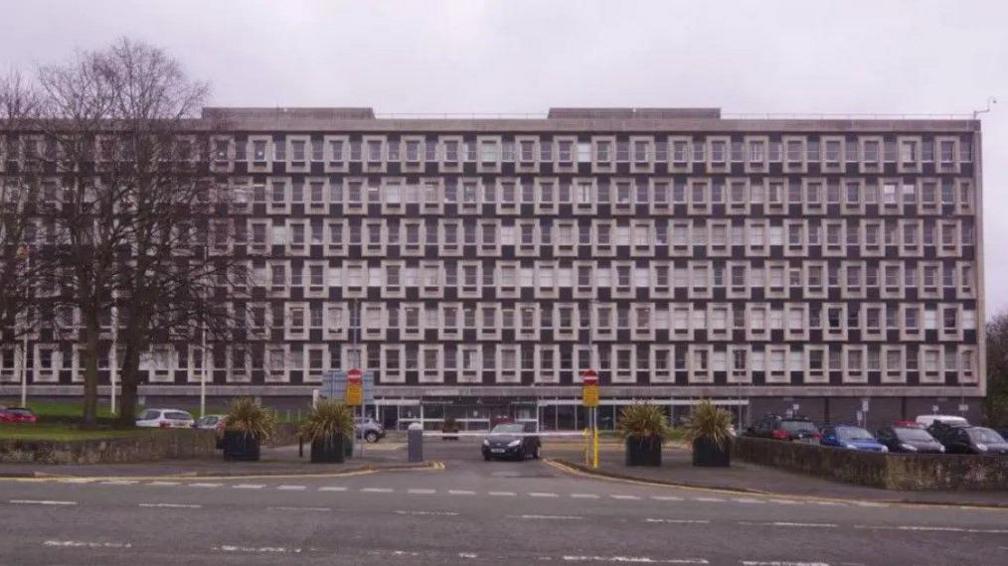 The exterior of county hall in Mold, which has seven floors and a black and white design. A car park with a barrier can be seen in front of the building.