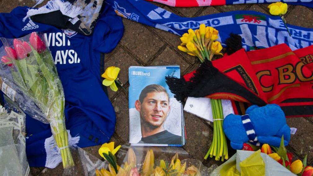 Flowers and football tops and scarves on the ground, surrounding a photograph of a young man, on the front of a magazine entitled "Bluebird".