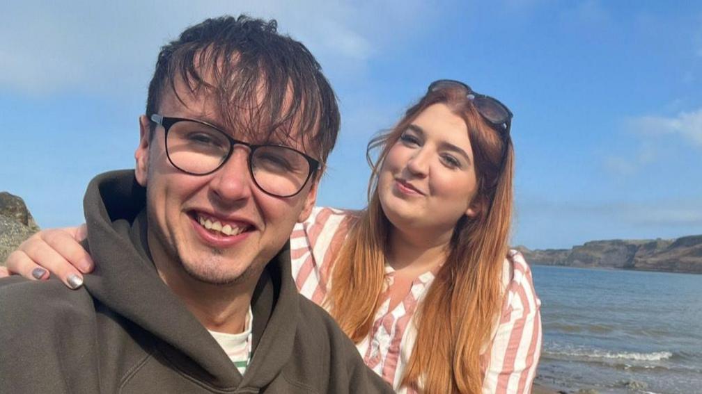 A young woman rests her hand on the shoulder of a young man with glasses on as both sit on a rocky beach with the sea disappearing behind them in the distance. It's a blue-sky, sunny day and both are smiling warmly.