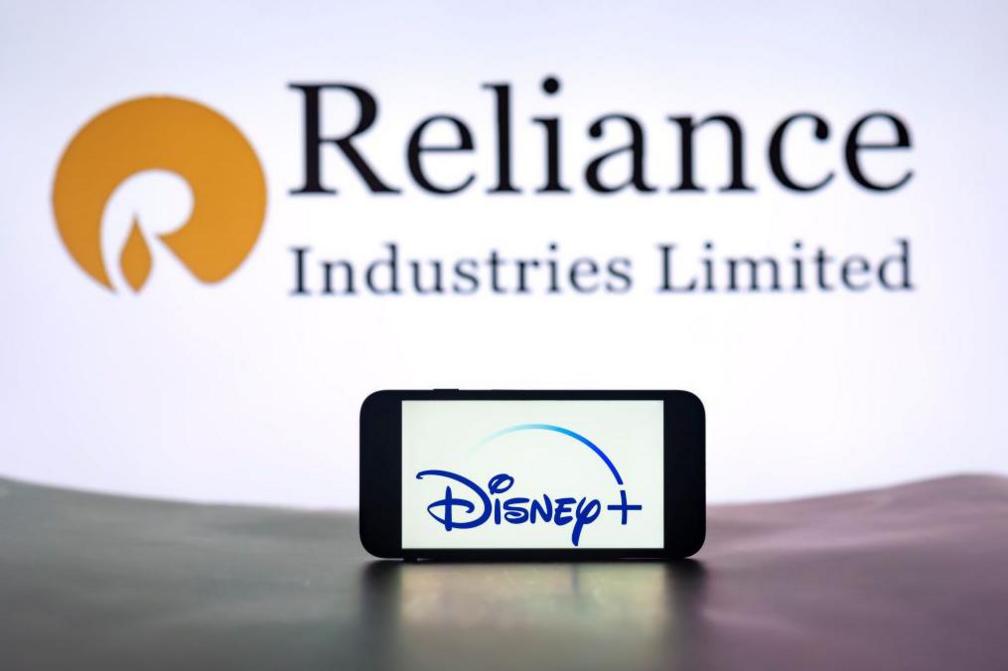  In this photo illustration, the Disney+ logo is seen displayed on a mobile phone screen with Reliance Industries Limited in the background