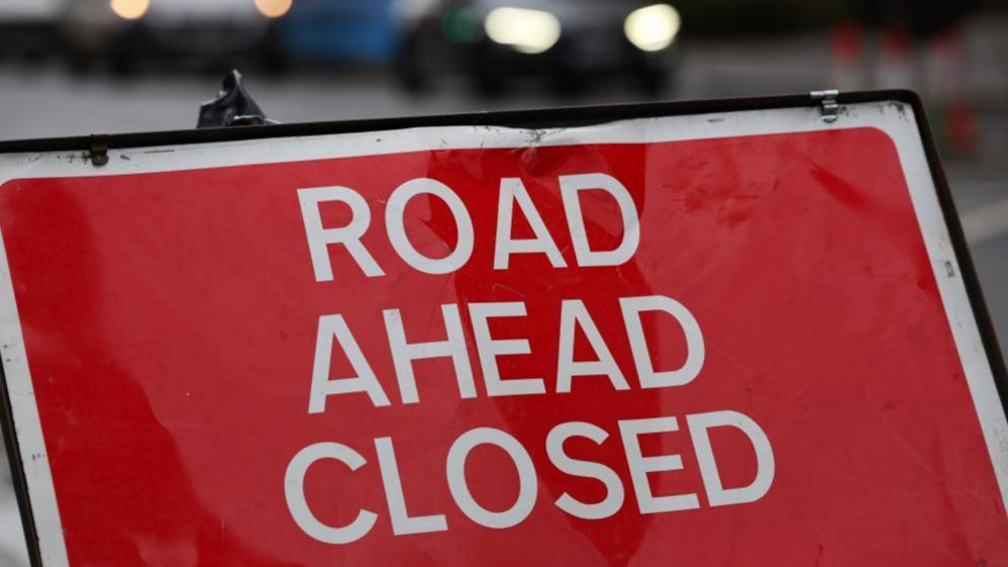 A close up image of a bright red road sign reading "Road Ahead Closed" is on a road. There are blurred cars behind the sign. 