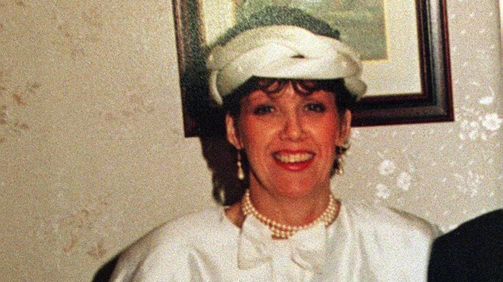 Geraldine Breslin wearing a white hat, a pearl necklace wrapped twice round her neck and a white dress. The wall behind here had white floral wallpaper and a picture with a dark wooden frame. 