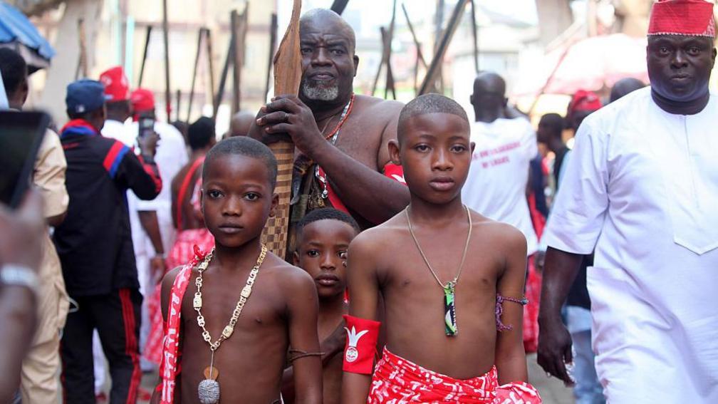 Young boys wearing red and white patterned traditional Yoruba clothes stand in front of men carrying what looks like sticks. Some of the men behind are wearing red and white clothing also - Wednesday 20 August 2025.
