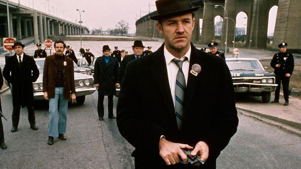 Policeman Gene Hackman is shown in a movie still walking toward the camera. He is holding a gun in his hands with policemen and police vehicles in the background