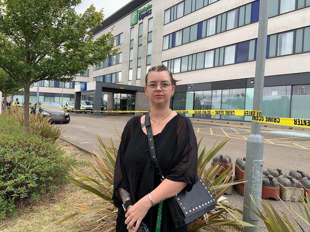 A woman with dark hair and glasses stands in front of yellow police tape in front of a large hotel building