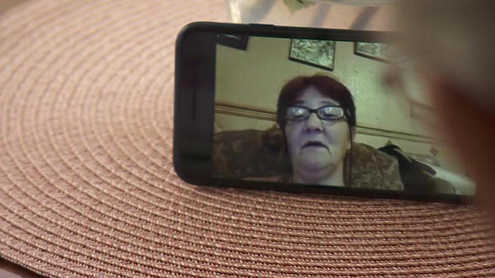 An image seen on a mobile phone of a woman wearing glasses sitting on a chair. The mobile is on a flat surface, being viewed by someone - their face is partially in the photo on the far right.