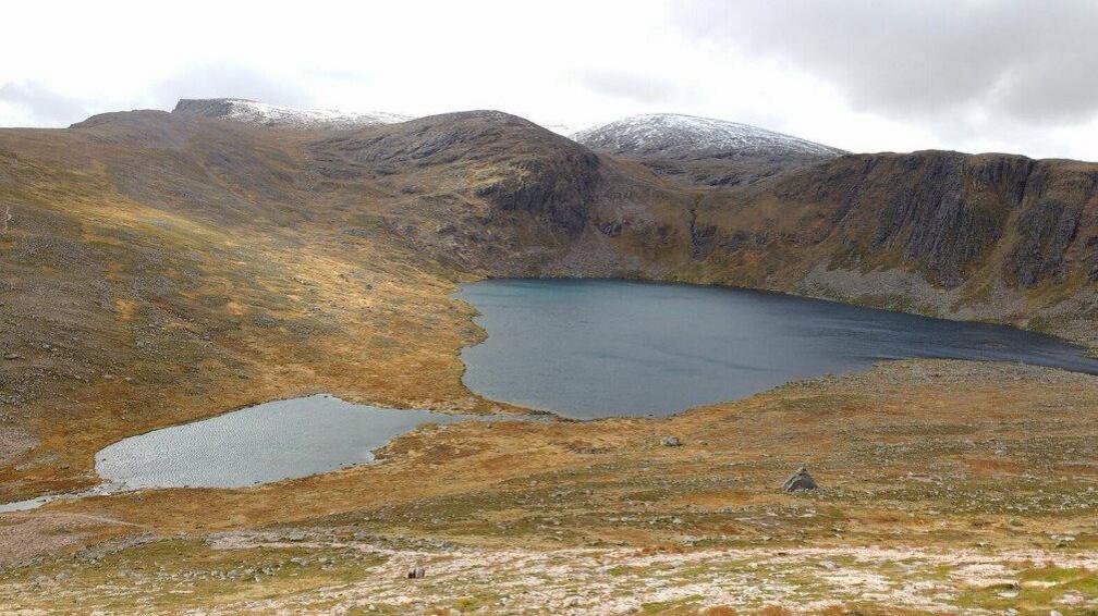 A dark lake surrounded by rocks, moors and peaks. The peaks furthest in the distance are covered in snow.