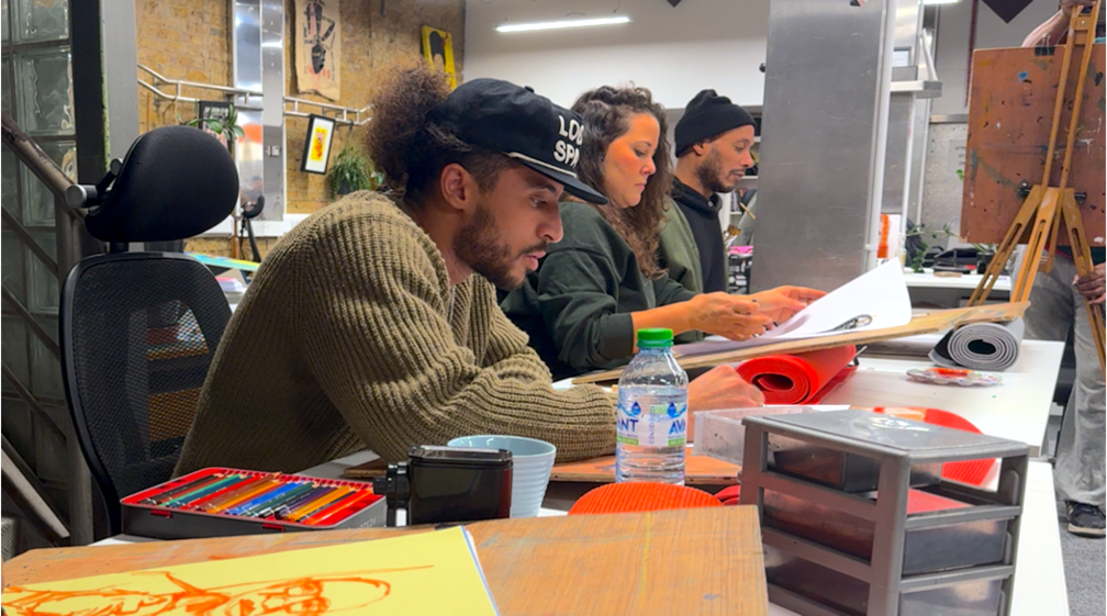 A man sits at a desk drawing a portrait. He is surrounded by art supplies and two people behind him are also drawing.