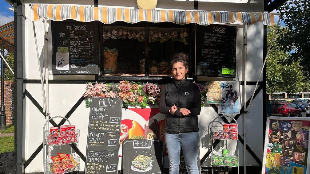 Keely Dean stands in front of her kiosk shop surrounded by products for sales and a chalkboard menu. She is wearing a black coat and blue jeans. The kiosk is bright and colourful with fake flowers below the counter. 