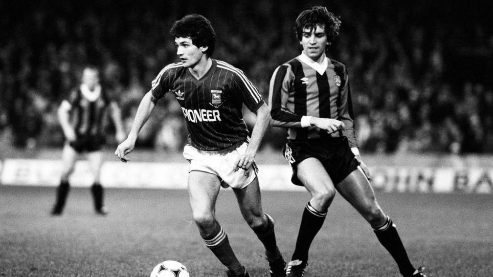 A black and white photo shows George Burley with a football at his feet. He's wearing a 1981 Ipswich home shirt with Pioneer sponsor. A Manchester City player looks on. A blurred crowd can be seen in the background