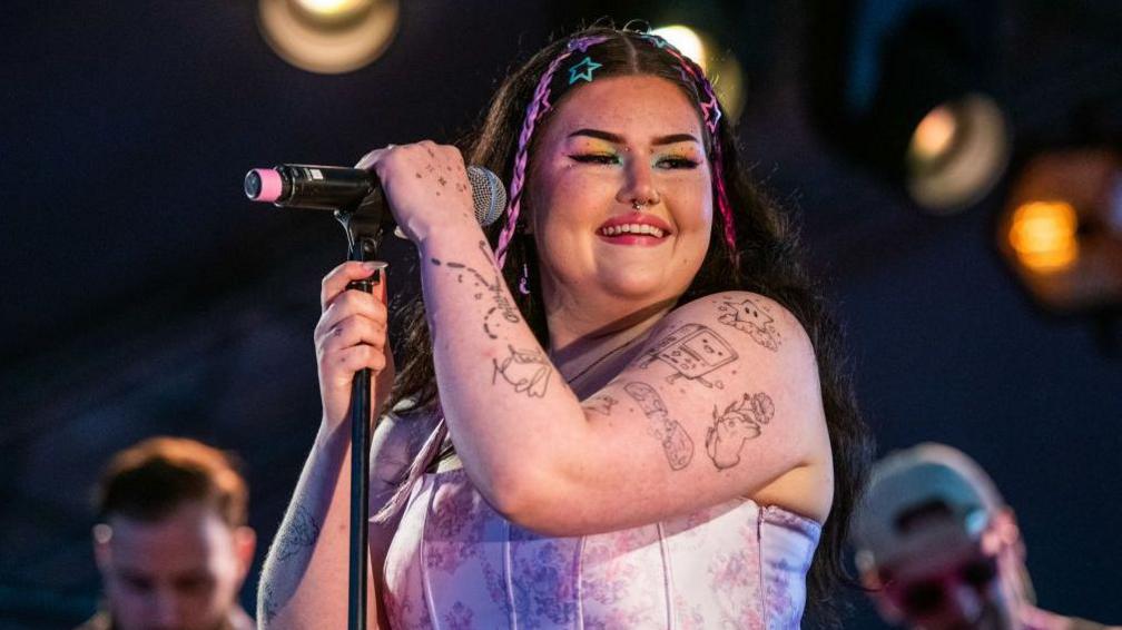 Mae Stephens performing, smiling, displaying her tattoos with colourful braids in her hair