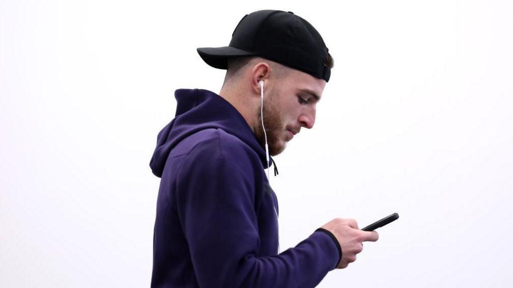 Declan Rice is looking down using his phone while wearing a backwards cap and a blue England tracksuit
