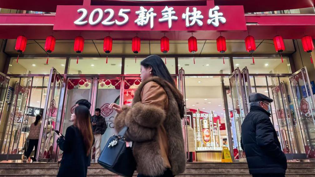 Pedestrians walk past a shopping mall decorated with red lanterns and a sign reading 2025 Happy New Year to celebrate the upcoming Chinese New Year on January 14, 2025 in Chongqing, China.