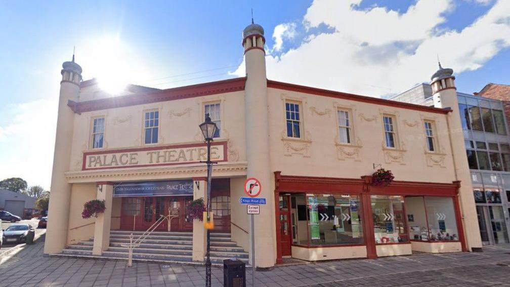A Google street view image of a cream-coloured, Neo-Byzantine-style building with deep red window and door frames. The entrance, which faces the corner of a street, has a sign reading 'Palace Theatre'  above two columns and steps leading to the doors.
