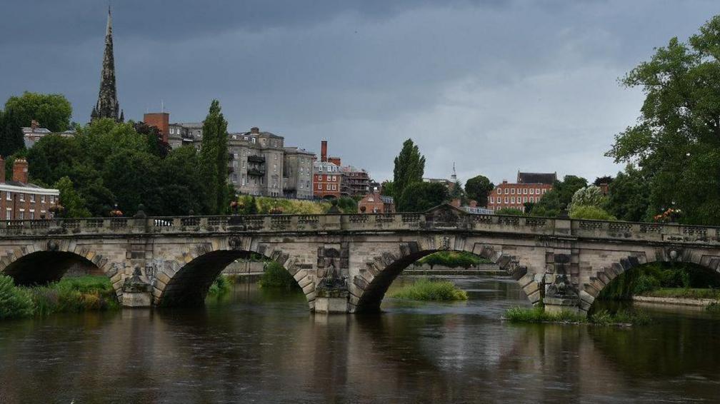 A stone bridge stands across a wide river, with a number of old buildings on the bank to the left, and all sat under dramatic grey skies 