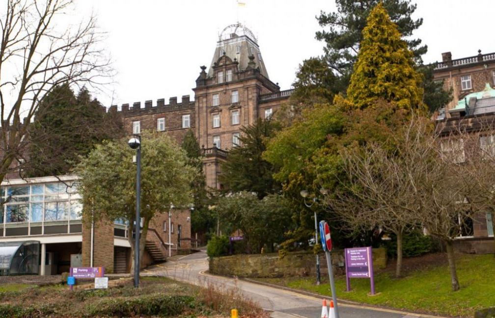 Grand council building set on a slope with a lower glass-fronted building. Two traffic cones in red and white are on the side of the roadway, which is lined by grass and medium trees, some which have lost their leaves. There are purple coloured signs with white lettering to direct visitors to the site.