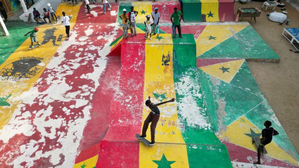 A high-angle wide shot of a bustling colourful skate park in the Ngor neighbourhood of Dakar, Senegal. The park is a vibrant hub of activity with skaters, either active on the ramps or stood watching. The entire concrete surface of the park, including curved transitions and steep ramps, is painted in the bold green, yellow and red colours of the Senegalese flag