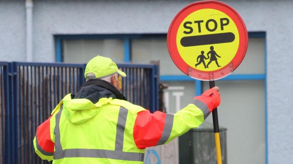 A school crossing patrol man in a yellow cap and high visibility jacket holds up a stop children sign