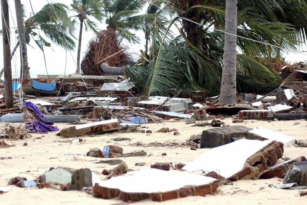 Debris and shattered structures litter the sandy coastline of Arugam Bay, with uprooted palm trees and remnants of buildings reflecting the devastation caused by the 2004 tsunami.