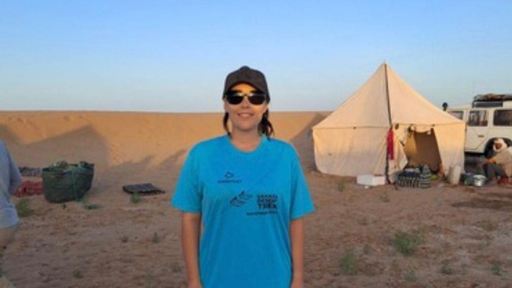 A woman in a cap, sunglasses and blue t-shirt standing in the desert with a tent in the background