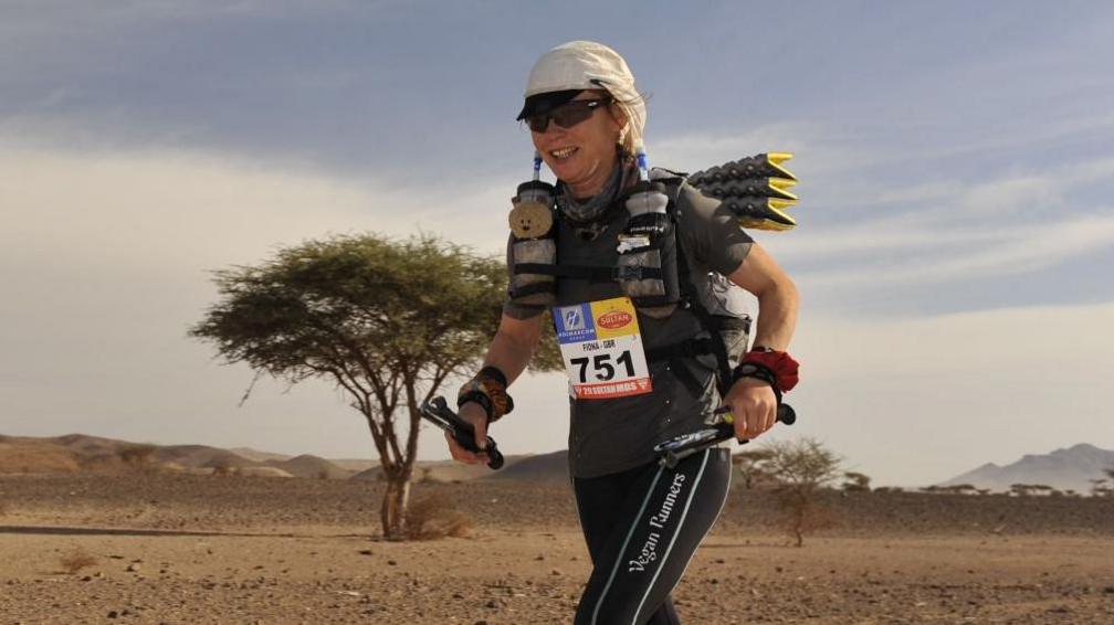 Fiona Oakes wearing her running gear while on a run. She is wearing a white hat and sports gear. She is in a desert landscape. It is cloudy above her.