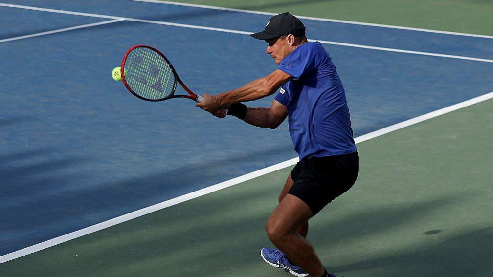 Kamil Majchrzak pictured wearing a blue t-shirt, black shorts and a black cap. He holds a tennis racket in his hand.