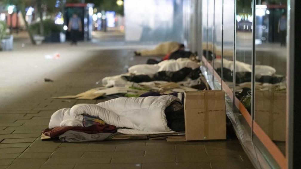 People in sleeping bags lie down on the pavement against a glass-fronted building