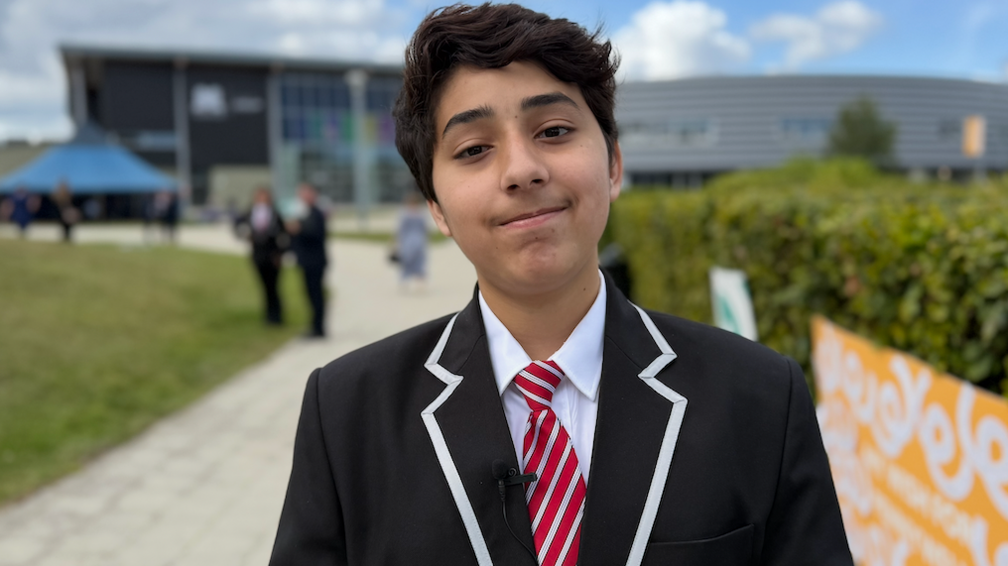 Salah is standing in front of school which is grey and blurred. He is smiling, has dark hair and is wearing a black blazer with a white trim and a red and white, striped tie. There are two other pupils in the background which is out of focus.