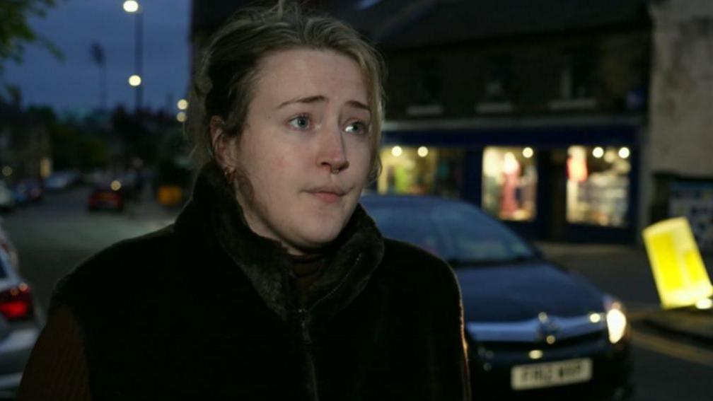 A woman stands next to a road, dressed in a dark coat. A car is just about to drive past her and can be seen over her left shoulder. Three illuminated shop fronts can be seen across the street.