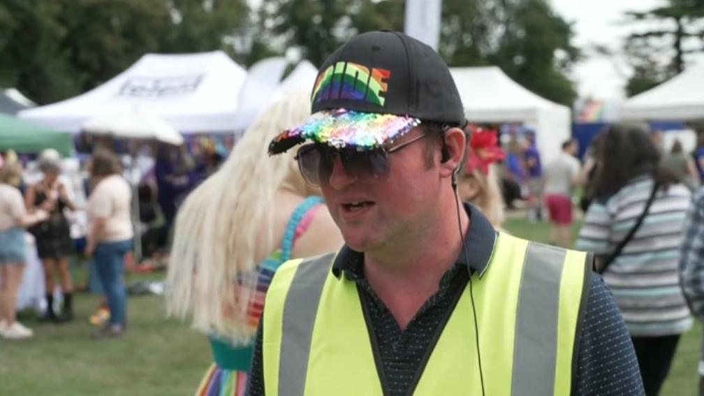 Daniel Browne, from Warwickshire Pride, wearing a cap with a sparkler front and the word "Pride" on it in rainbow colours. He has a high-vis jacket on over a blue dotted shirt with festival-goers behind him