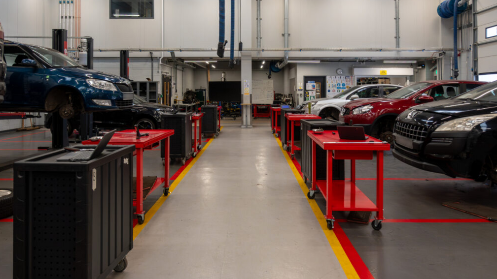 Two rows of cars can be seen on the left and right of the image. They are parked in bays with some raised up as if being worked on. There is a gap in between the two rows marked off with red and yellow paint.