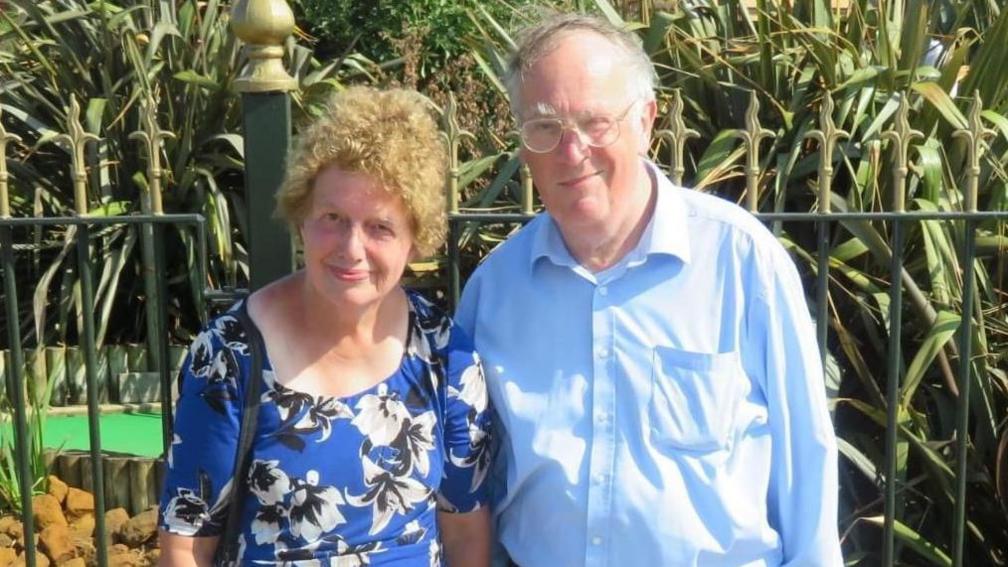 Lois and John McCullough standing in front of a mini golf course. They are both wearing blue and smiling at the camera.