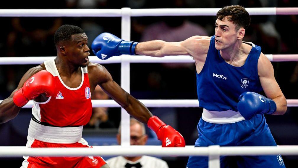 Two men are boxing in a ring. The man on the left is black and dressed in red and white kit and gloves, the man on the right is white and dressed in blue and white kit and gloves. The background is out of focus, but there is an audience behind them and a male referee in a white shirt and black bow tie. 