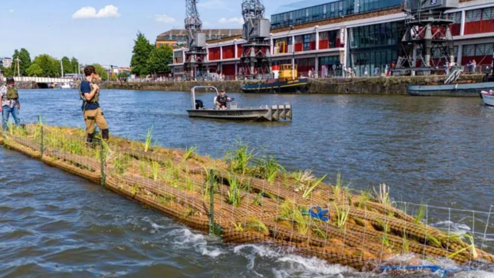 Men stood on recycled plastic water pipes in the middle of the water in front  of the Mshed.