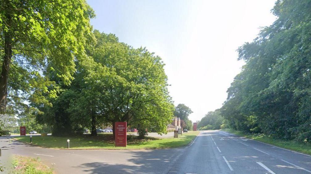 Streetview image of A614 at Clumber Park Hotel, showing a single carriageway road with a turning lane, and hotel buildings behind trees. 