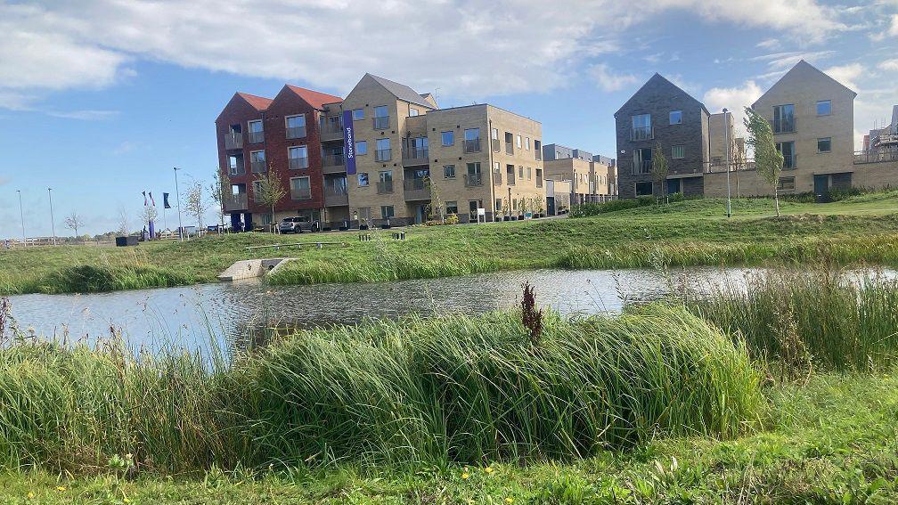 A close up of a lake and green field, behind which stand a number of new three and four-story houses. 