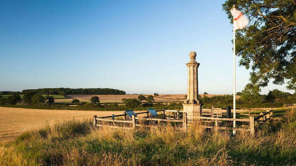 A stone monument encased in a wooden fence in the middle of a field. A flag pole with the red and white flag of England atop it is next to it. 