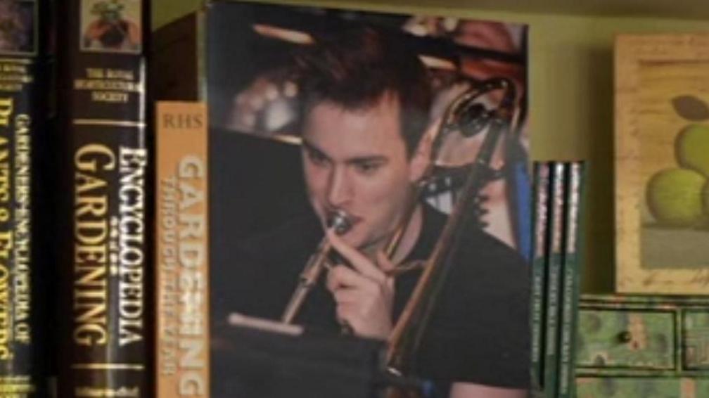A printed photograph of a young man with brown hair, playing the trumpet, sits between books on a shelf.