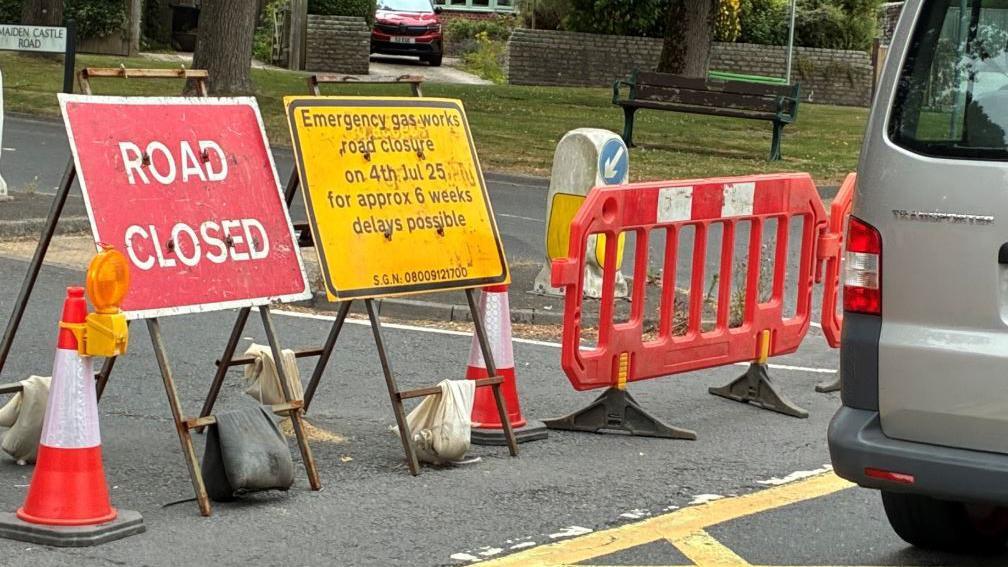 A red road closed sign and a yellow sign stating: "Emergency gas works road closure on 4th Jul 25 for approx 6 weeks, delays possible", can be seen at the end of Maiden Castle Road. There is also a red barrier and cones.