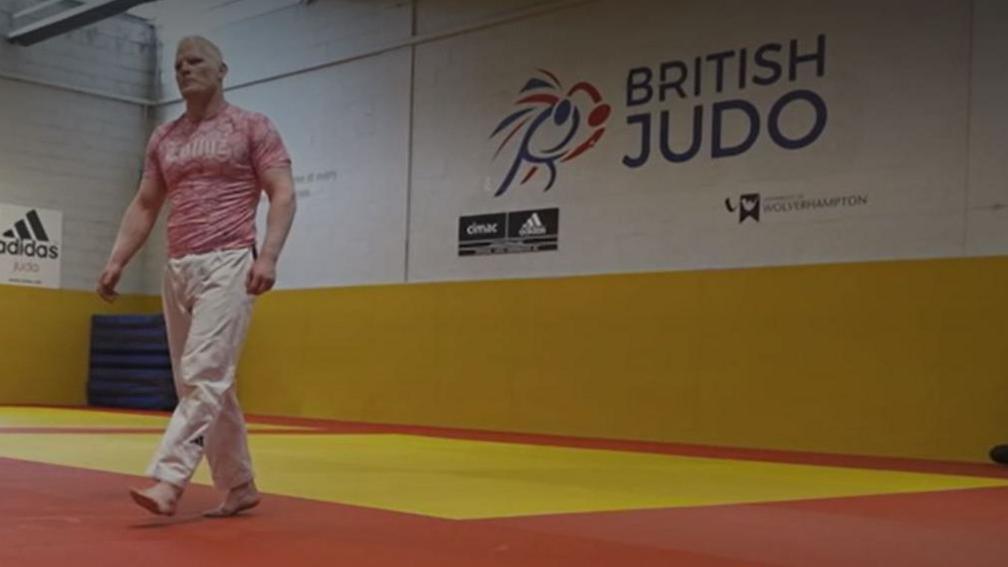 Mr Molloy walks across a sports hall with British Judo logo on the wall