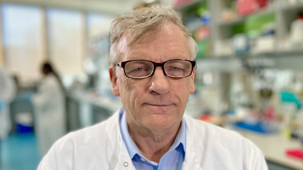 A head and shoulders image of Prof Colin Cooper wearing a white lab coat and blue shirt, with a blurred background of a scientific lab 