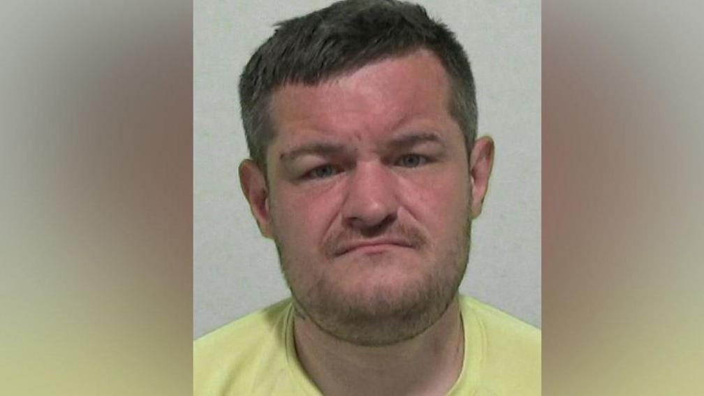 Mugshot of Trott who has short dark hair and a thin wispy stubble beard, he is wearing a bright yellow T-shirt.